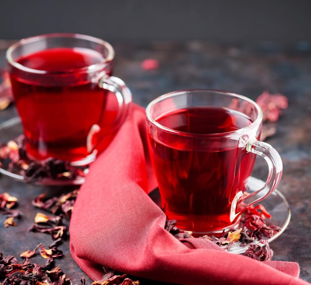 two glass mug filled with red hibiscus tea with dried hibiscus petals scattered around them